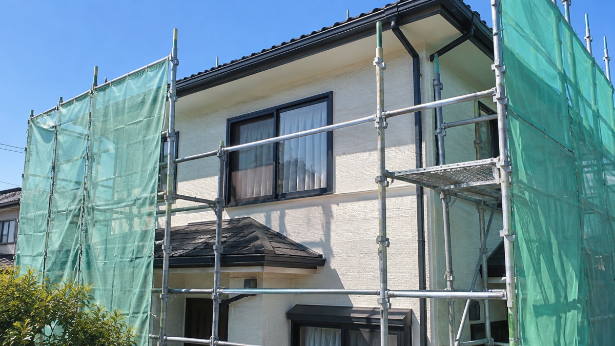 足場と緑色の防護シートに囲まれた、クリーム色の外壁の2階建て住宅。晴れた空の下で、窓と瓦屋根が見える