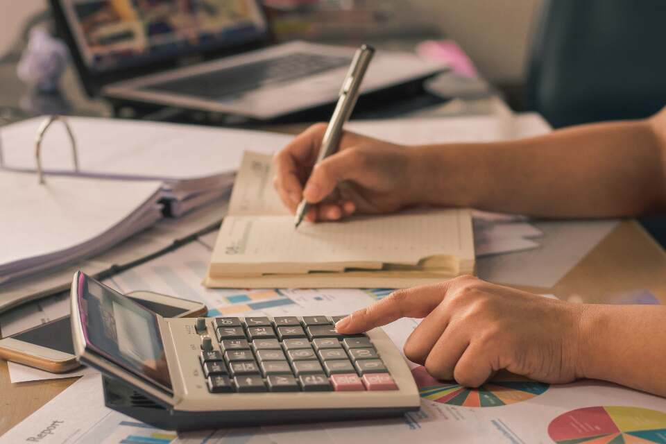 電卓を押す手と、ノートにペンで書き込む手。机にはファイルとグラフの資料が広がっている