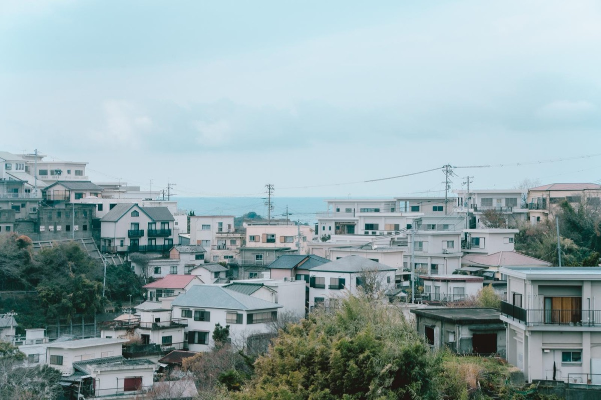 海が見える斜面に、白やベージュの外壁の住宅が重なって並ぶ。空はうすい曇りで、手前に木々がある